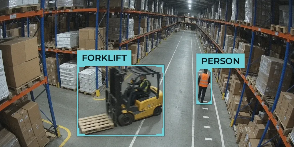 CCTV view of a warehouse racking aisle with a forklift carrying an empty pallet and a worker in hi-vis walking behind