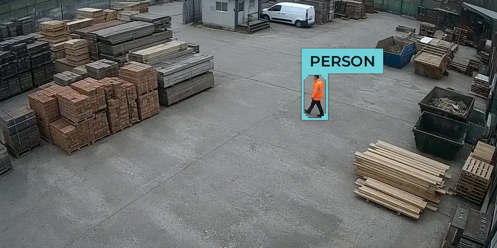 CCTV view of a builders merchant yard with one worker in an orange hi-vis jacket walking between stacks of bricks and timber