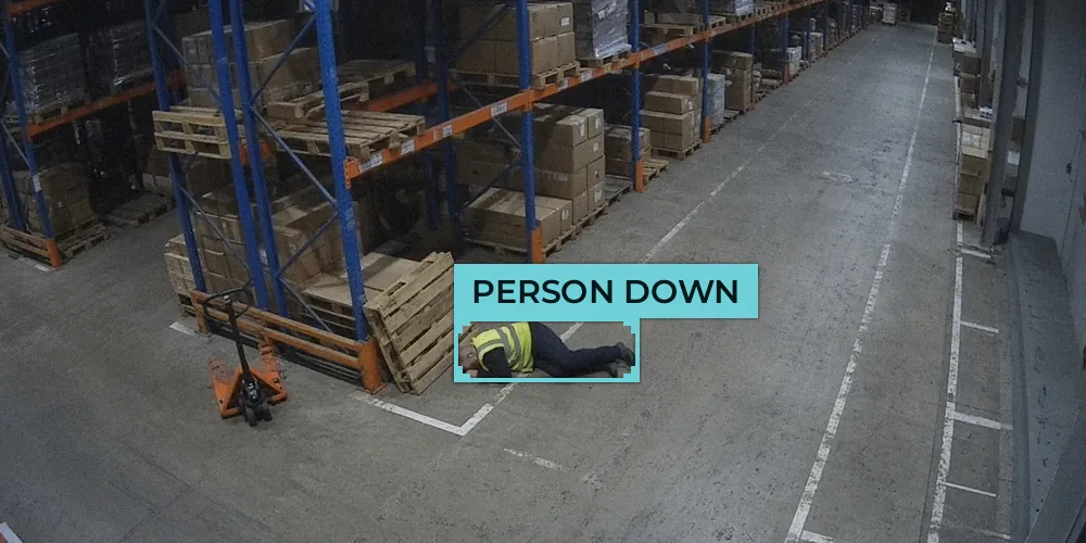 CCTV view of a warehouse aisle with a worker in a hi-vis vest lying on the floor beside a pallet