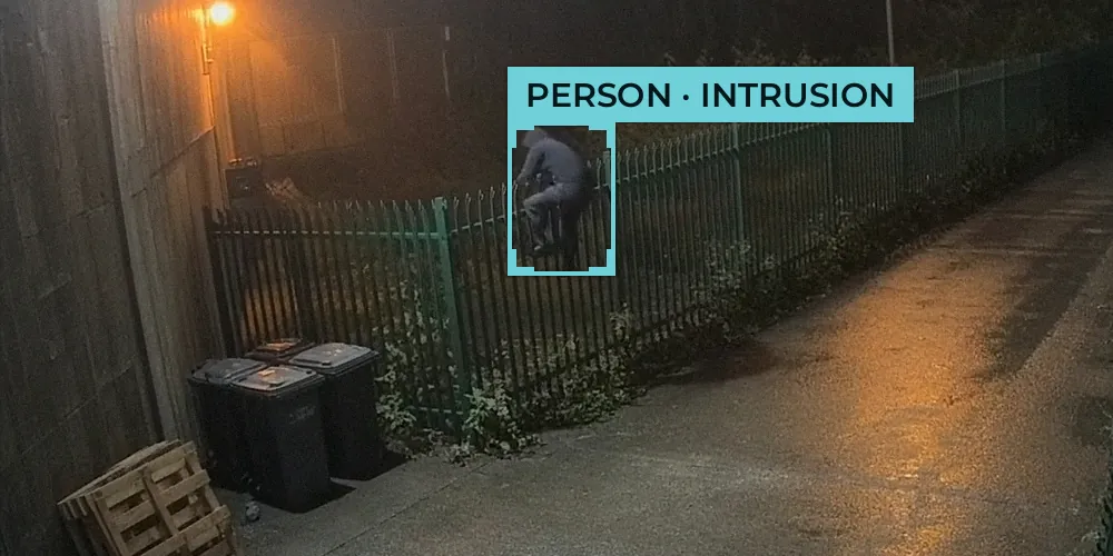 Night-time CCTV view of a person in a dark hooded top climbing a green palisade fence beside wheelie bins
