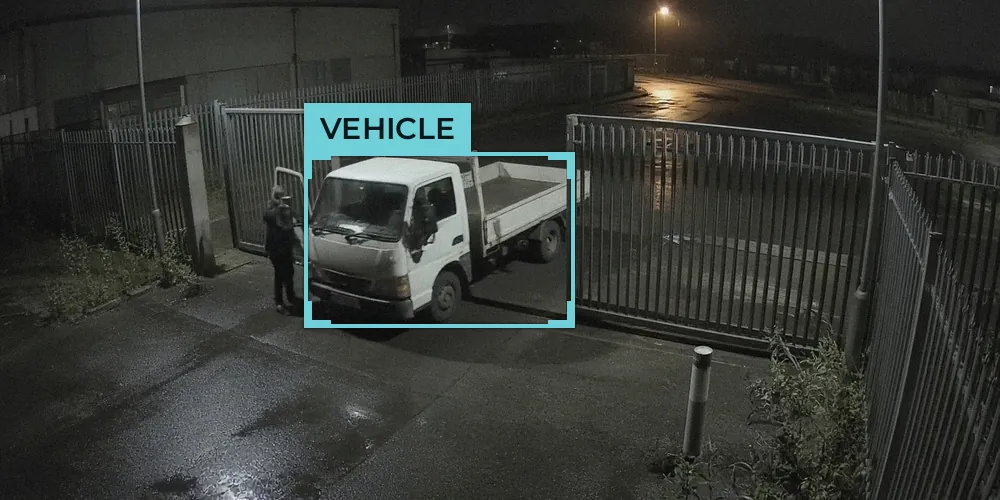 Night-time CCTV view of a white flatbed truck stopped in an open steel gate with a person beside the driver door