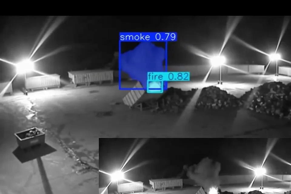 Real night-time infrared CCTV frame of a storage yard with smoke and a small fire beside a skip, each boxed and labelled by Woodcock AI