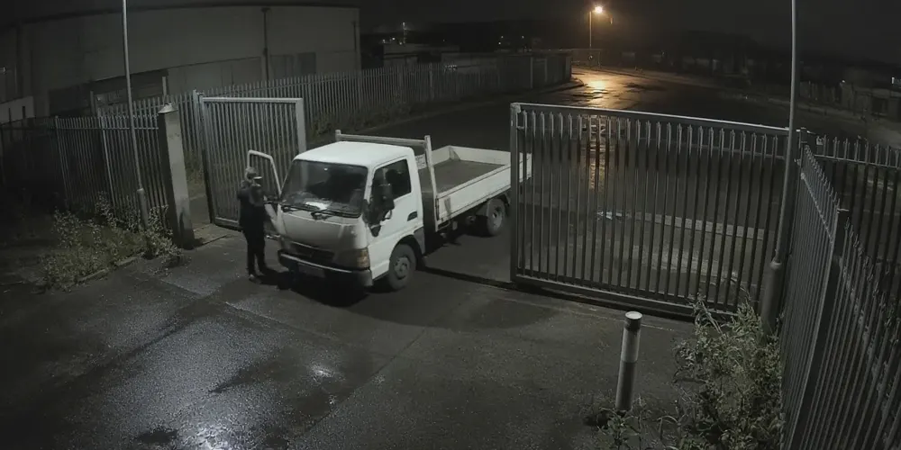 Illustrative night-time CCTV frame of a site gate with a person standing beside a pickup truck